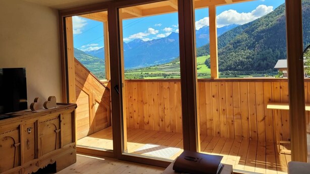 A room with a wooden floor, a television, and a large window with a mountain view.