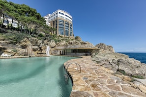Una piscina con plataforma de piedra, cascada y un edificio con grandes ventanales al fondo.