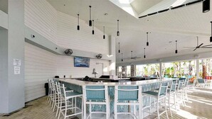 A bar area with white bar stools and a long counter, surrounded by a high ceiling with hanging lights and a fan. - Brand New Home at Windsor Cay Resort with Lazy River (Clermont)