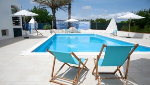 Ein Swimmingpool mit zwei blauen Stühlen und Sonnenschirmen, umgeben von einem weißen Gebäude und Palmen.