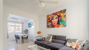 A modern living room with a grey sofa, a dining area with chairs, a glass coffee table, a ceiling fan, and a large painting on the wall. - Holiday Home 'Horizon Oasis' with Sea View, Wi-Fi and Air Conditioning (Santa Pola)