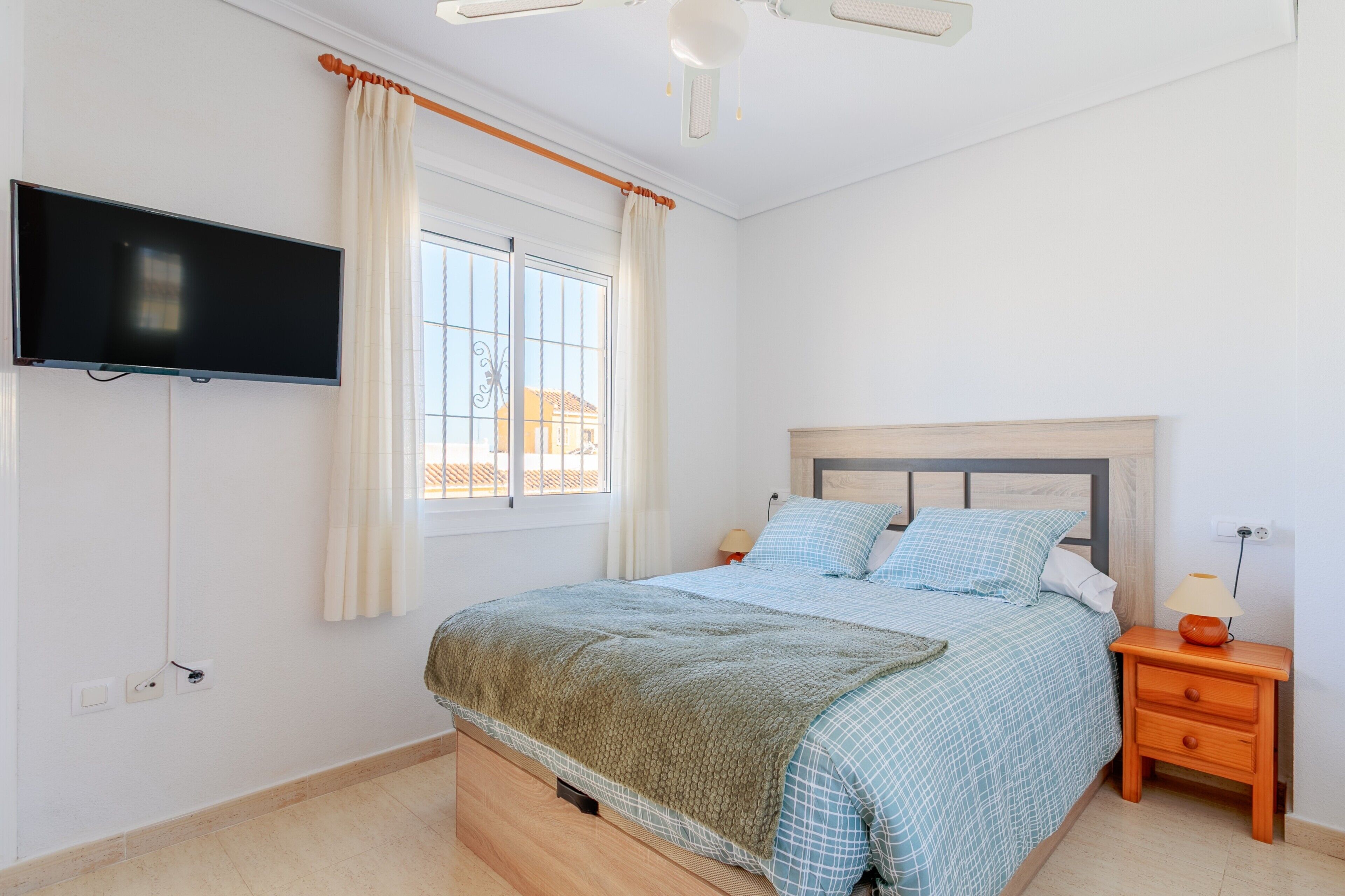 Een slaapkamer met een bed, een aan de muur bevestigde televisie, een raam met gordijnen, en een nachtkastje met een lamp.