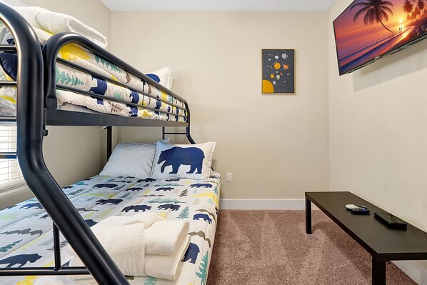 A bunk bed with a bear-themed bedding, a small table, a wall-mounted TV, and a framed picture of a sunset. - Reunion Village | Discover Florida's Hidden Gem (Kissimmee)