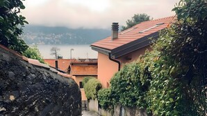 Un chemin de pierre mène à un bâtiment dont le toit est recouvert de tuiles rouges. Il est entouré de verdure et offre une vue sur une étendue d’eau.