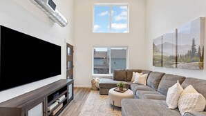 Uma sala de estar moderna com uma grande televisão de tela plana, um sofá seccional, uma mesa de centro e uma janela grande com vista para uma casa.
