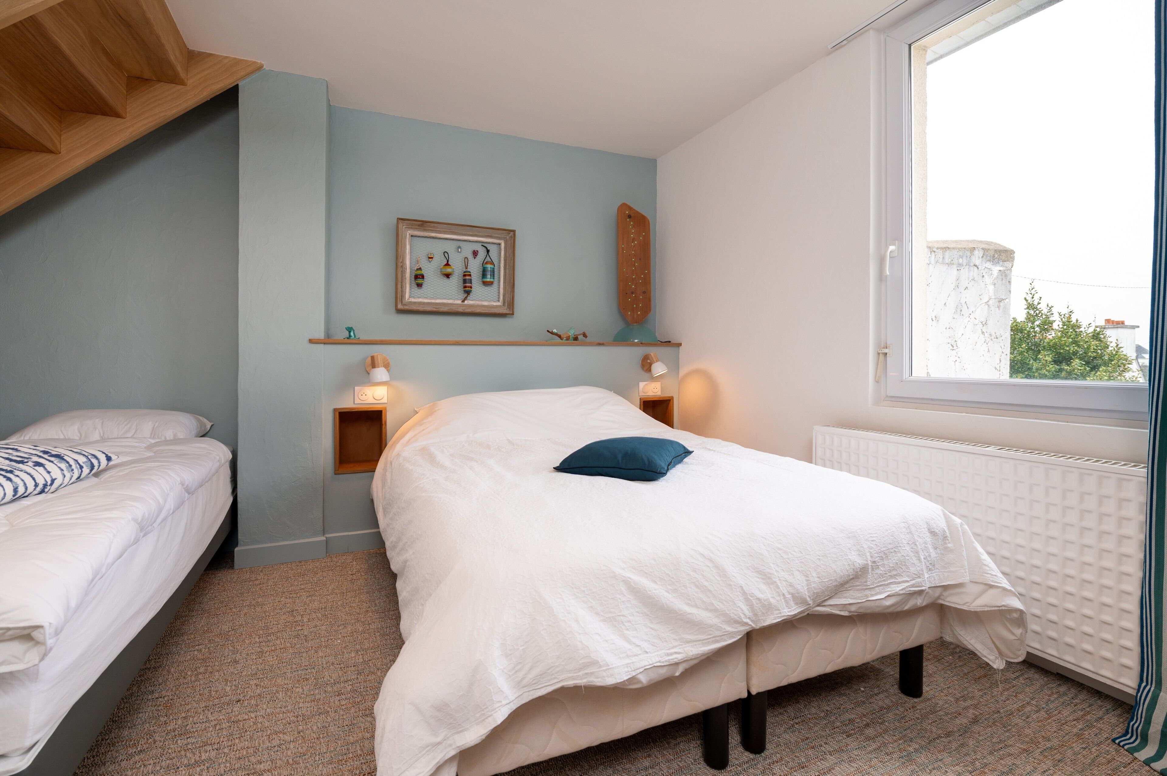 Een slaapkamer met twee bedden, een raam met uitzicht, een plank met decoratieve spullen, en een aan de muur hangend schilderij.