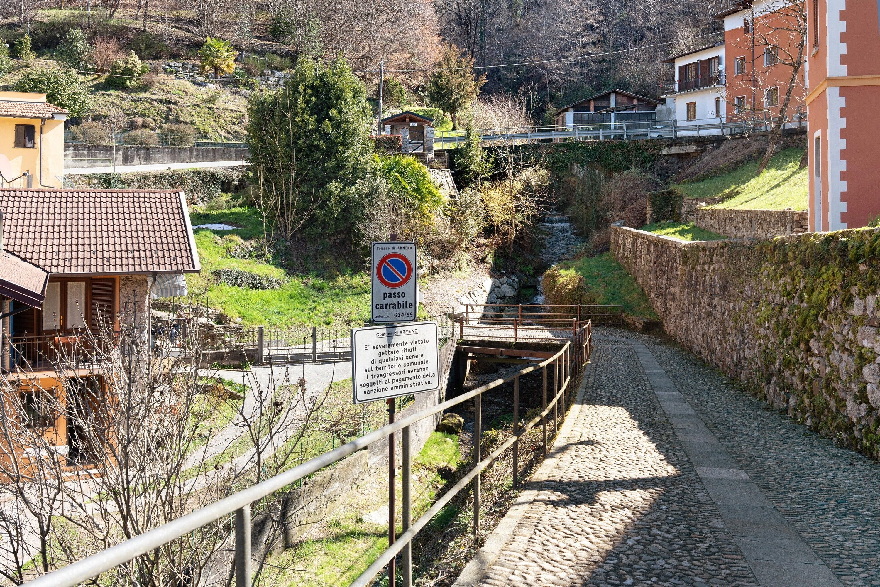 Un étroit sentier pavé de pierres menant à un pont enjambant une rivière, le tout entouré de maisons et de verdure.