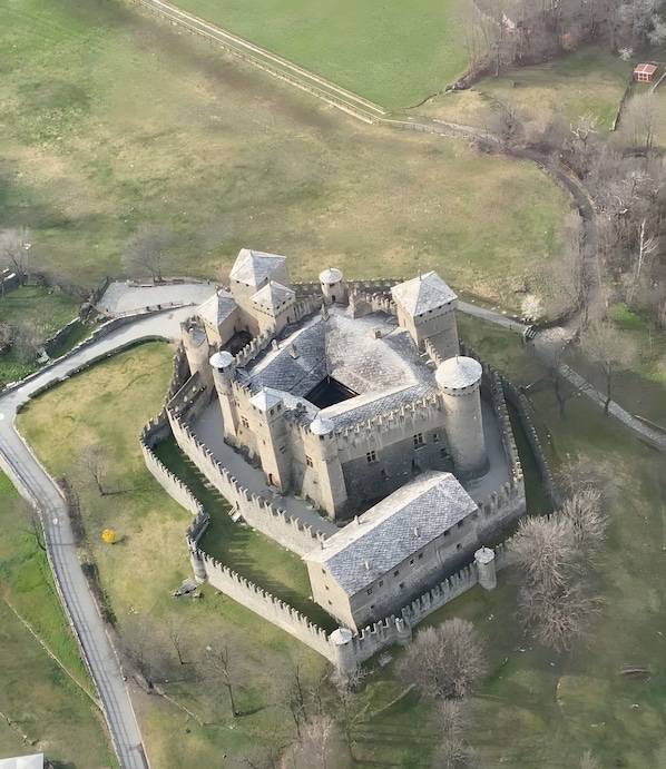 An aerial view of a historic castle with a moat, surrounded by grassy fields and a paved road.