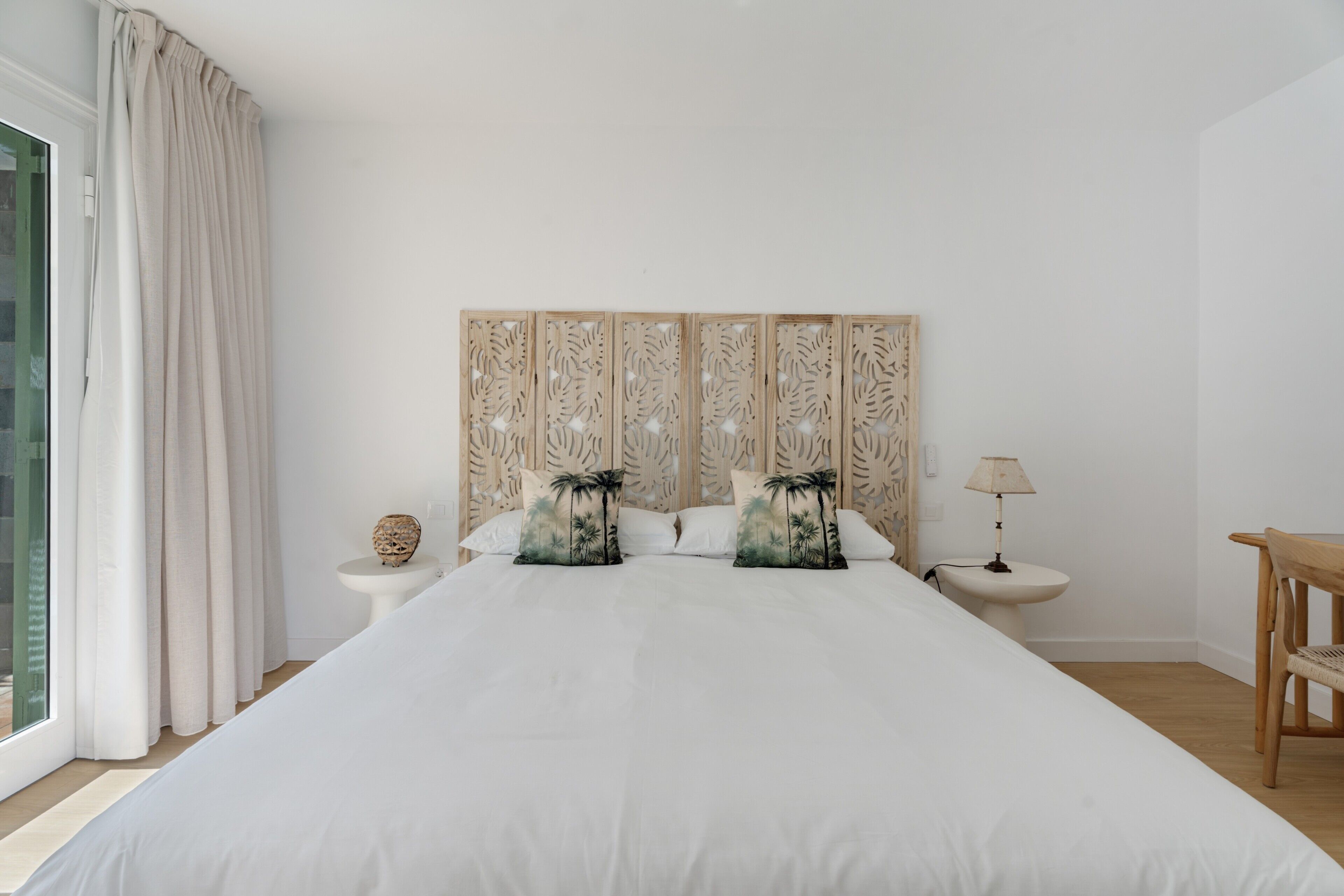 Un dormitorio con una cama grande, dos mesitas de noche, un cabecero de madera, una ventana con cortinas y una pequeña mesa.