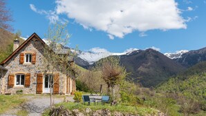 Una cabaña de piedra con chimenea de piedra, contraventanas de madera y una mesa con sillas en un paisaje montañoso.
