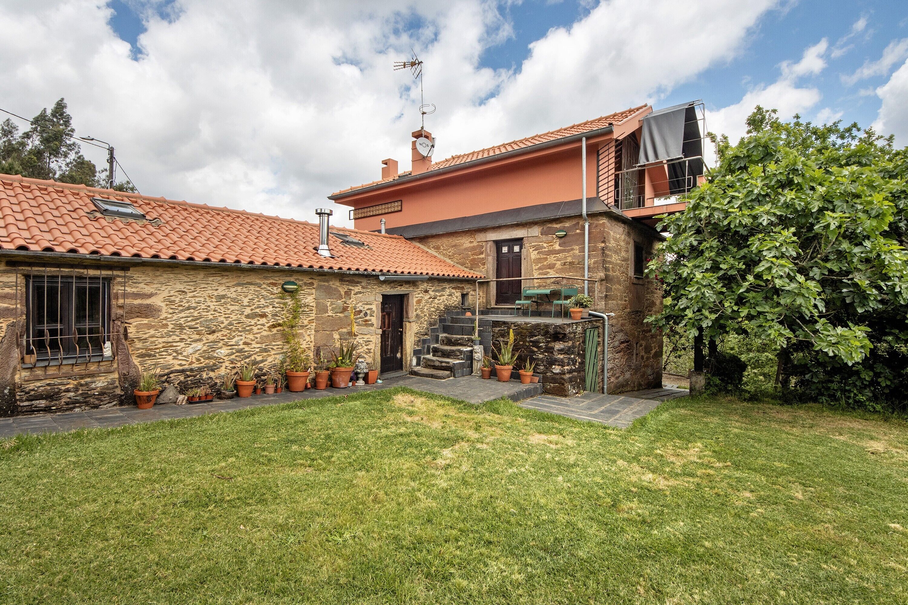 A stone house with a red-tiled roof, a small garden, and a staircase leading to a balcony.