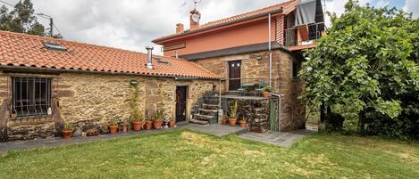 A stone house with a red-tiled roof, a small garden, and a staircase leading to a balcony.