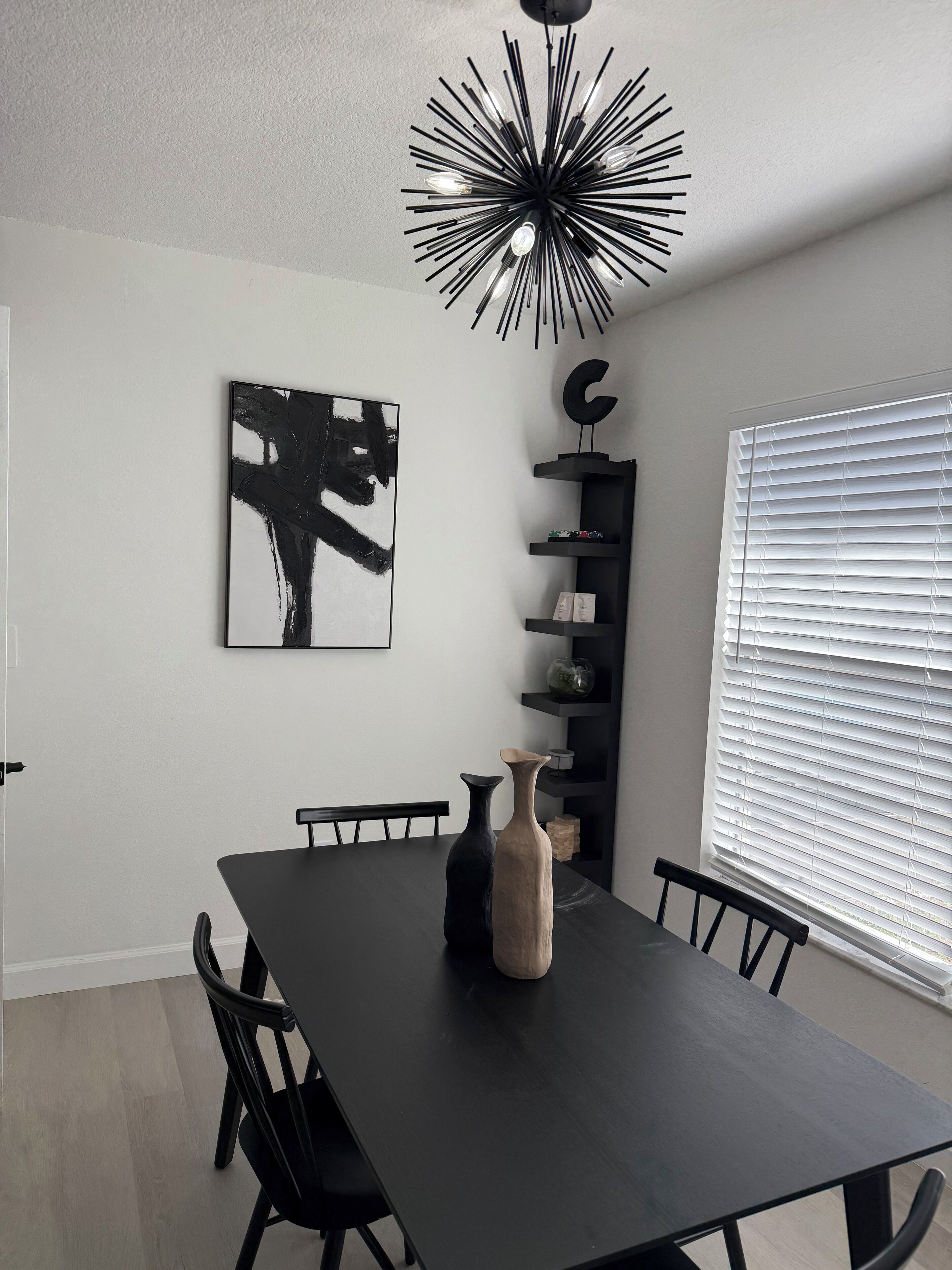 A dining area with a black table, black chairs, a black vase, a black abstract painting, a black ceiling light fixture, and a window with blinds.
