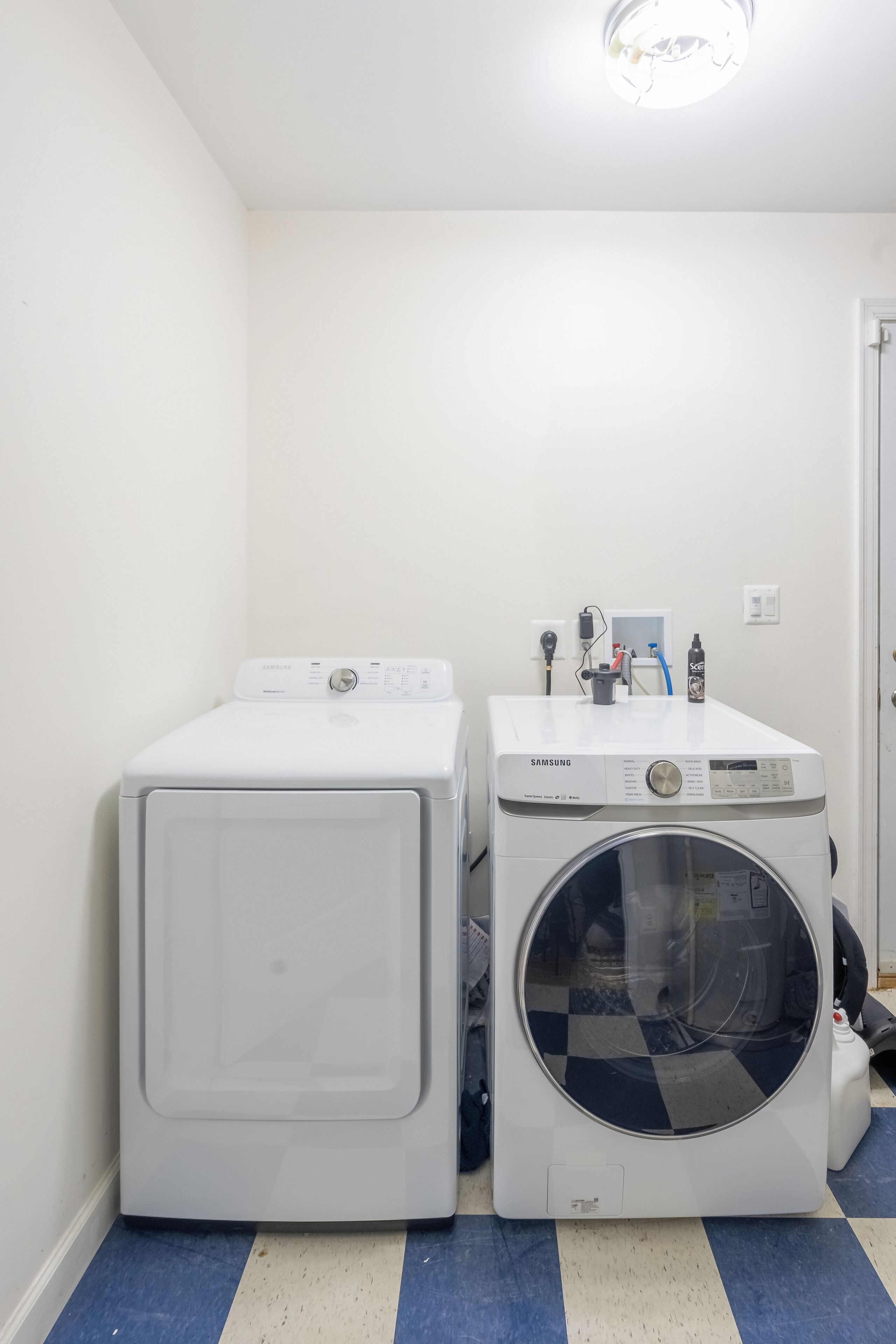 A laundry room with two white washing machines and a dryer, a blue and white checkered floor, and a white door in the background.
