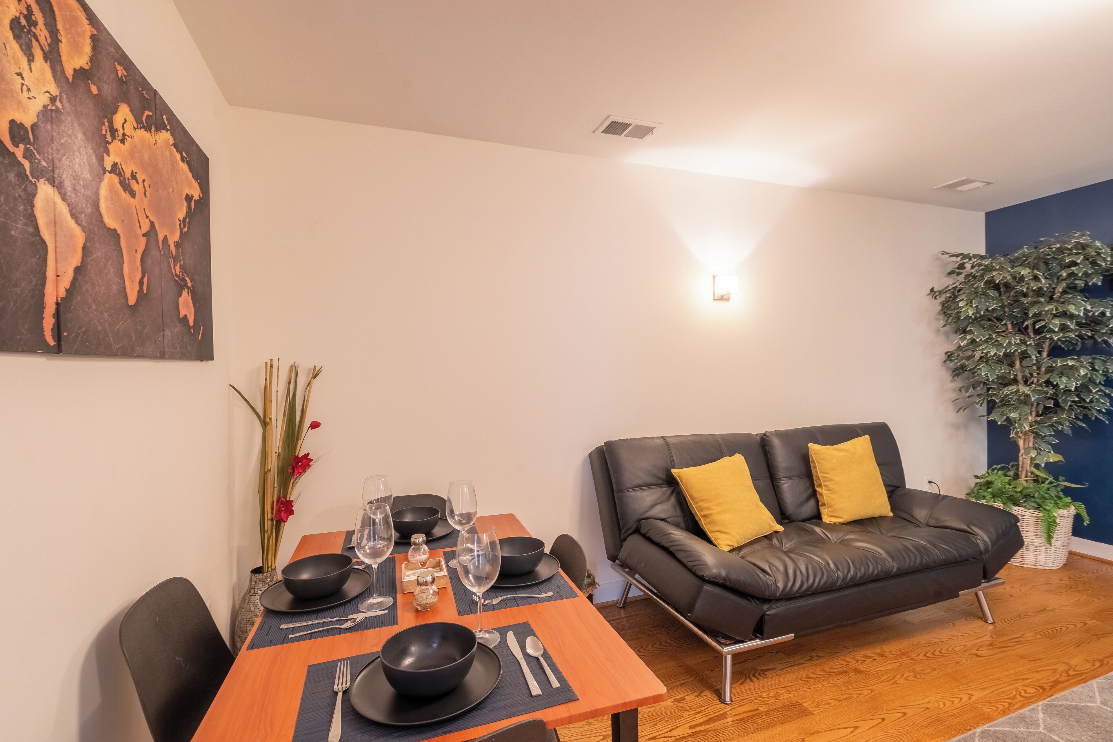 Un coin repas avec une table en bois dressée pour quatre personnes, des bols noirs, des verres à vin et des couverts. Un canapé en cuir noir avec des coussins jaunes est placé contre un mur bleu, et une grande plante se trouve dans un coin.