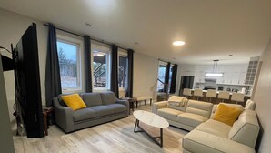 A modern living room with a grey sofa, a beige sofa, and a coffee table. There is a kitchen area with white cabinets and a dining table with chairs.