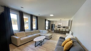 A modern living room with a sectional sofa, a grey sofa with yellow cushions, a small round table, and a kitchen area with a bar counter and appliances.