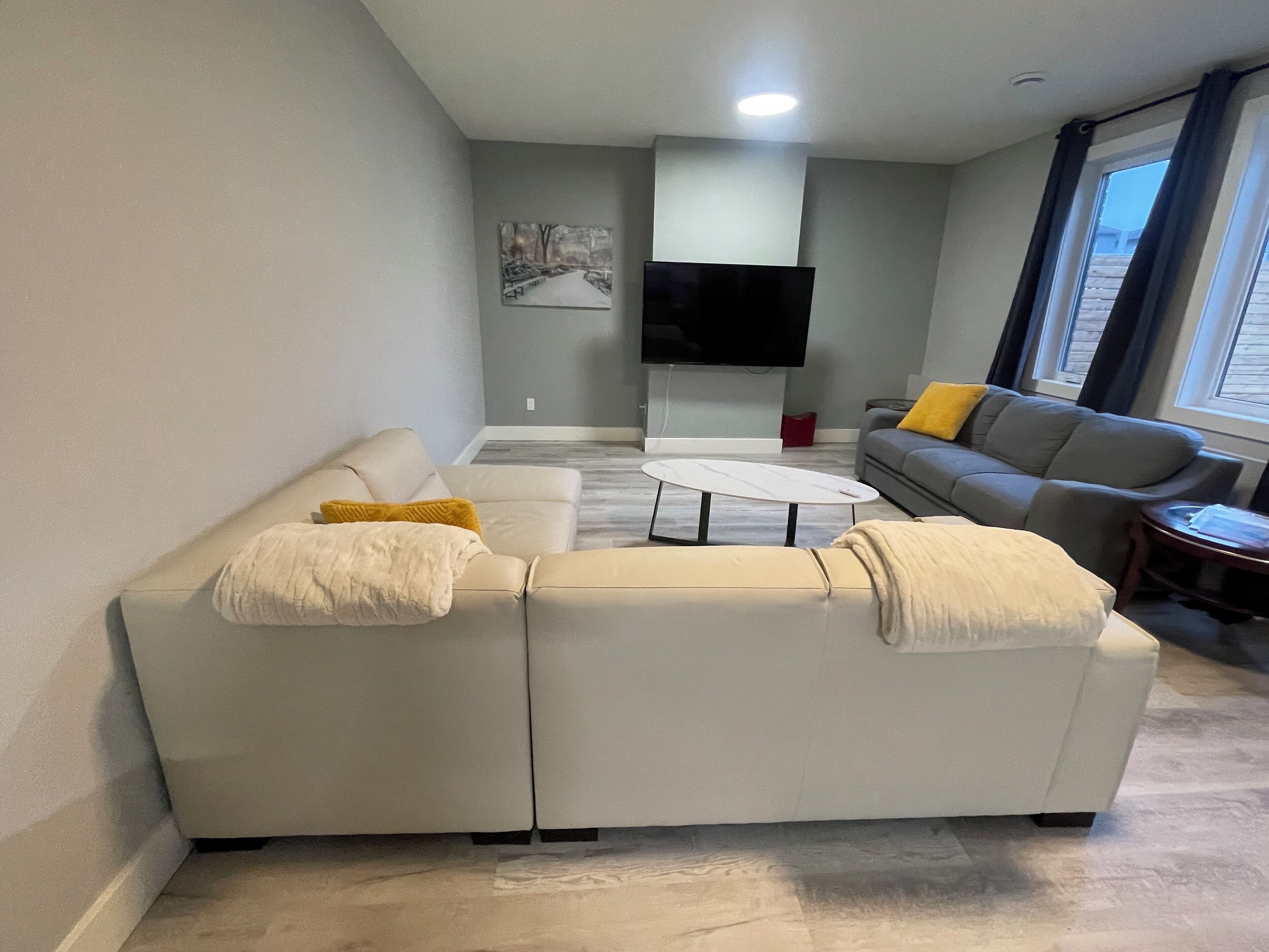 A modern living room with a sectional sofa, a coffee table, and a grey sofa with a yellow pillow. A flat-screen TV is mounted on the wall, and there is a window with curtains.