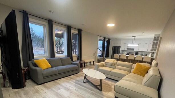 A modern living room with a grey sofa, a beige sofa, and a white coffee table. There are large windows with curtains, a staircase leading to a balcony, and a kitchen area in the background.