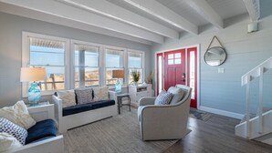 A living room with a sofa, armchairs, a coffee table, and a rug. There are large windows, a red door, and a staircase. - Pinstripe Paradise-Kitty Hawk MP 5, Ocean View, 10 Yds to Beach, Pool, Hot Tub, Pets OK, Flex Stays (Kitty Hawk)