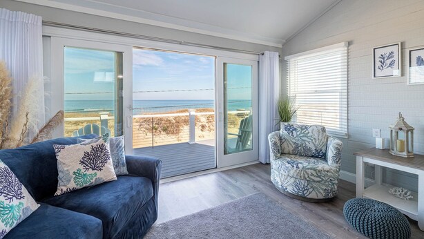 A living room with a view of the beach, a blue sofa, a patterned armchair, and a small table with a lantern. - Pinstripe Paradise-Kitty Hawk MP 5, Ocean View, 10 Yds to Beach, Pool, Hot Tub, Pets OK, Flex Stays (Kitty Hawk)