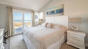 A bedroom with a large bed, a nightstand, a lamp, a painting, and a view of the beach. - Pinstripe Paradise-Kitty Hawk MP 5, Ocean View, 10 Yds to Beach, Pool, Hot Tub, Pets OK, Flex Stays (Kitty Hawk)