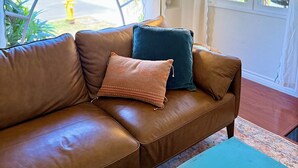 A brown leather sofa with two cushions, a blue table, and a window with a view of greenery and a yellow fire hydrant outside. - Charming, Historic 2-bedroom house in Coronado…28+ day discount (Coronado)