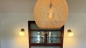 A wooden dining table with a central wooden bowl, surrounded by placemats with a pattern of small shapes. Above the table, there is a woven pendant light fixture. The background features a dark wooden cabinet with glass doors and two wall-mounted lights. - Charming, Historic 2-bedroom house in Coronado…28+ day discount (Coronado)