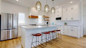 A modern kitchen with white cabinetry, a stainless steel refrigerator, a large island with bar stools, and pendant lighting. - Pet Friendly Beach Home, Shared Pool, Boardwalk to the Beach, Golf Cart Included (Port Aransas)