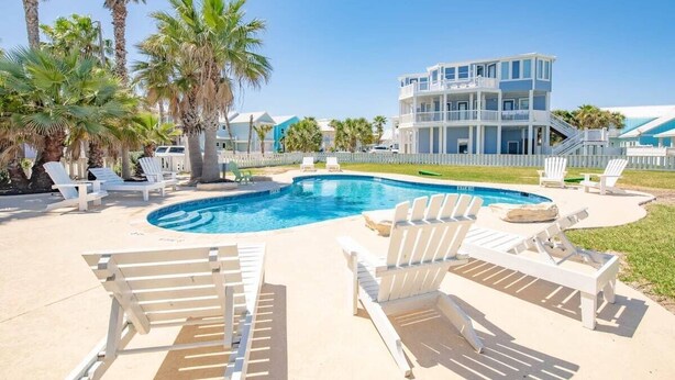 A pool area with lounge chairs, a large building, and palm trees. - Pet Friendly Beach Home, Shared Pool, Boardwalk to the Beach, Golf Cart Included (Port Aransas)
