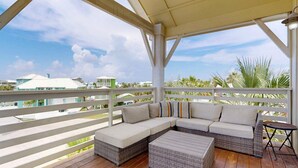 A covered outdoor seating area with a sofa and a small table, overlooking a residential area with houses and palm trees. - Pet Friendly Beach Home, Shared Pool, Boardwalk to the Beach, Golf Cart Included (Port Aransas)