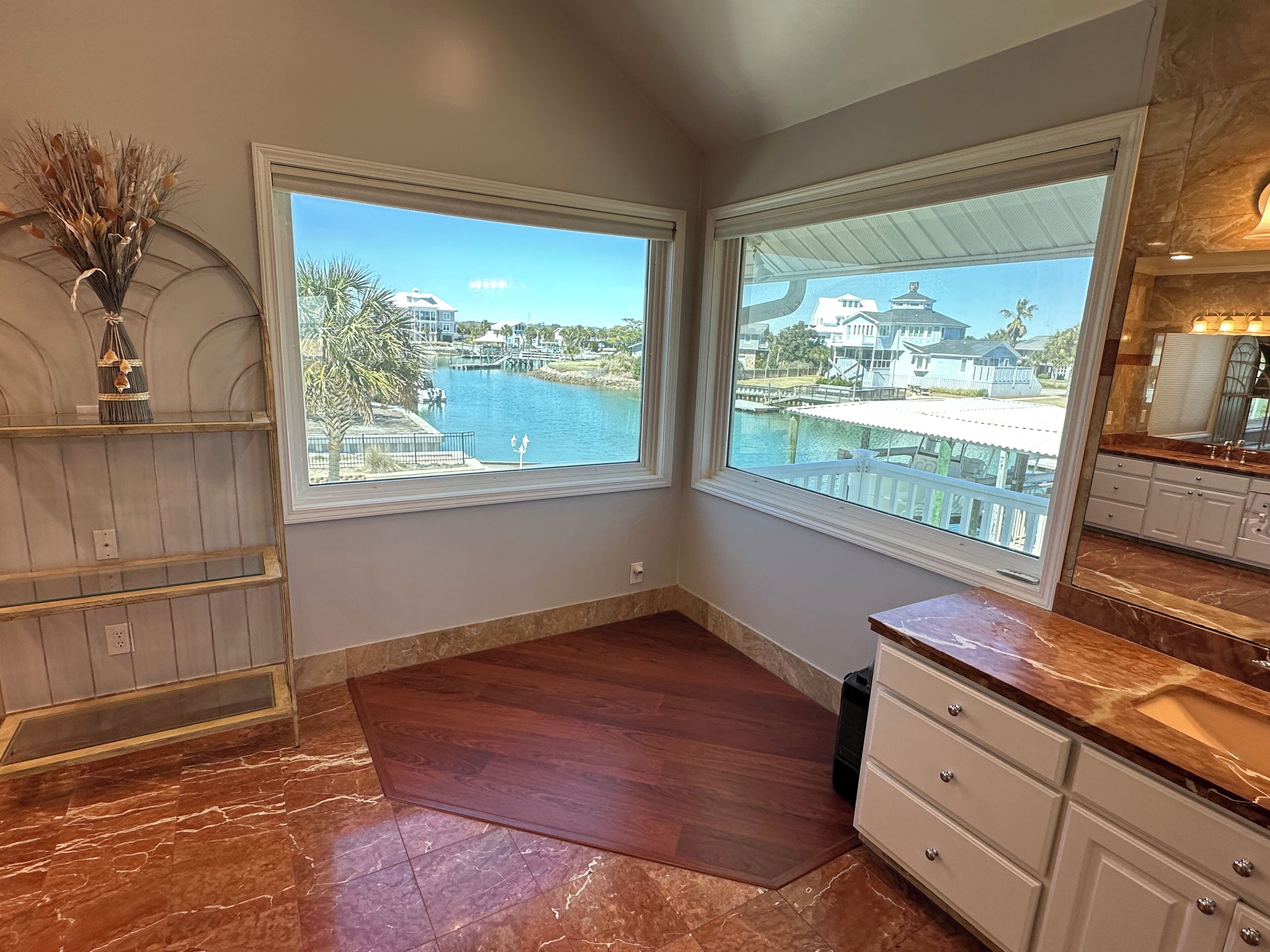 A corner room with a large window overlooking a body of water and a dock, wooden flooring, and a marble backsplash in the kitchen area.