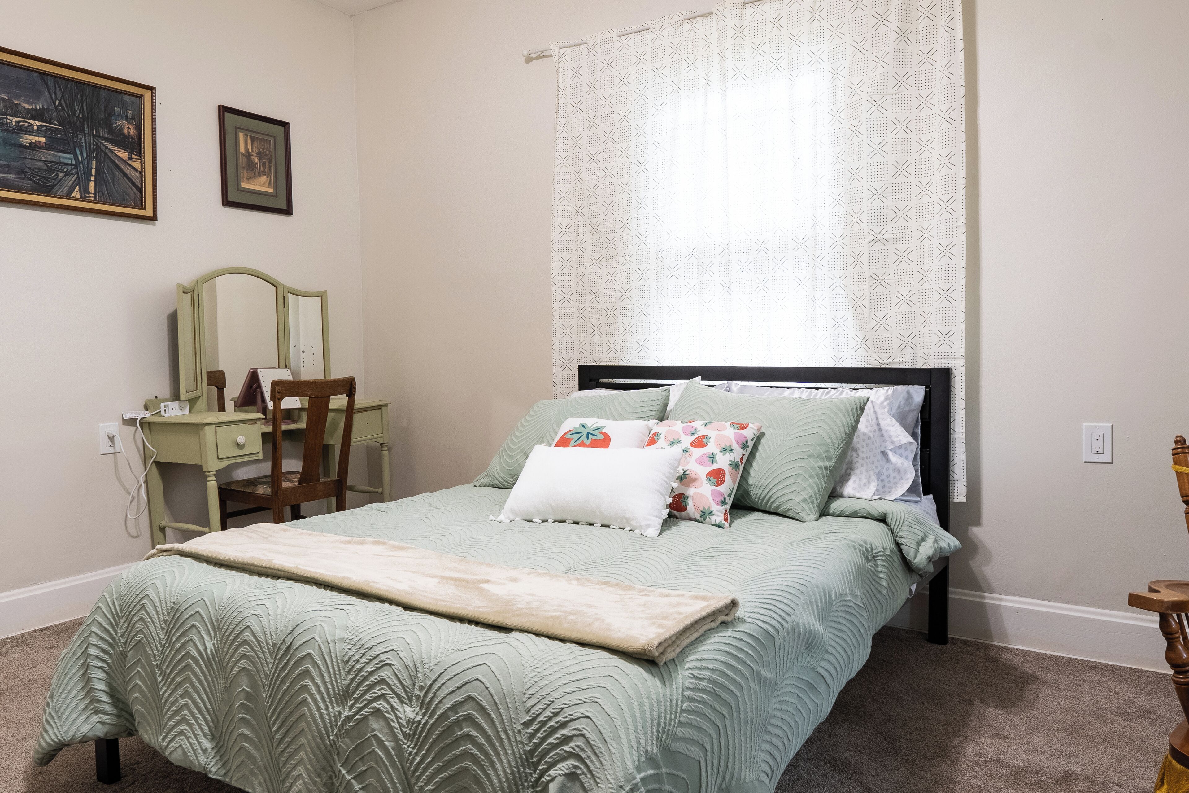 A bedroom with a bed, a desk, a chair, and a window with patterned curtains.