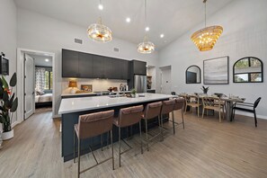 Een moderne keuken met donkere kastjes, een groot eiland met barkrukken, hanglampen en een zithoek met een tafel en stoelen.