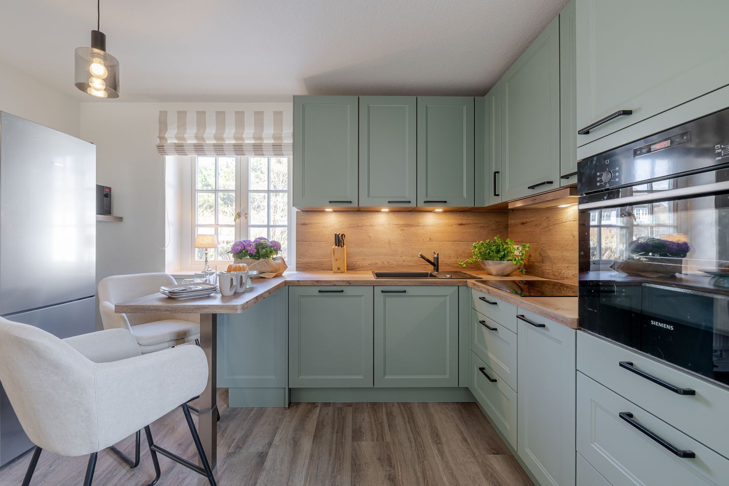 A modern kitchen with light green cabinets, a wooden countertop, a built-in oven, and a dining area with a white chair.