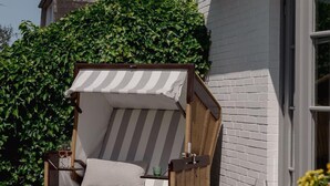 A patio with a striped canopy chair, a table set with glasses and fruit, and a white brick building in the background. - Baileys Hüs - Lovingly renovated "Baileys Hüs" in the picturesque village of Keitum on (Keitum)