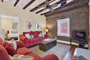 A living room with a red sofa, a wooden coffee table, and a television. There is a brick wall and a ceiling fan.