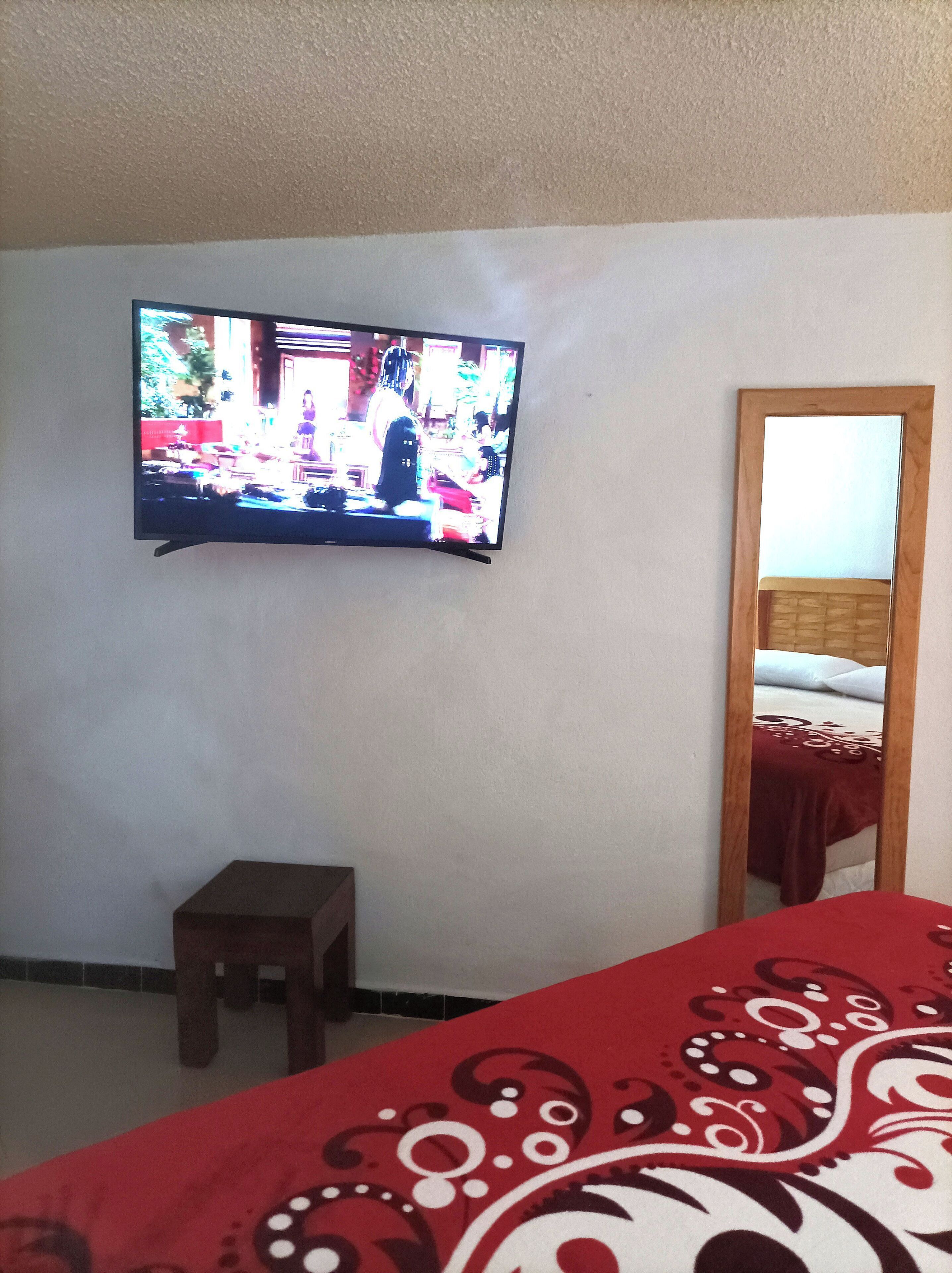 Un televisore montato a parete, uno sgabello di legno, un letto con copriletto rosso a motivi e uno specchio con cornice di legno.
