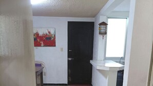 Un couloir avec un sol carrelé, une porte noire, une petite table avec une chaise violette, un lavabo blanc et une fenêtre.