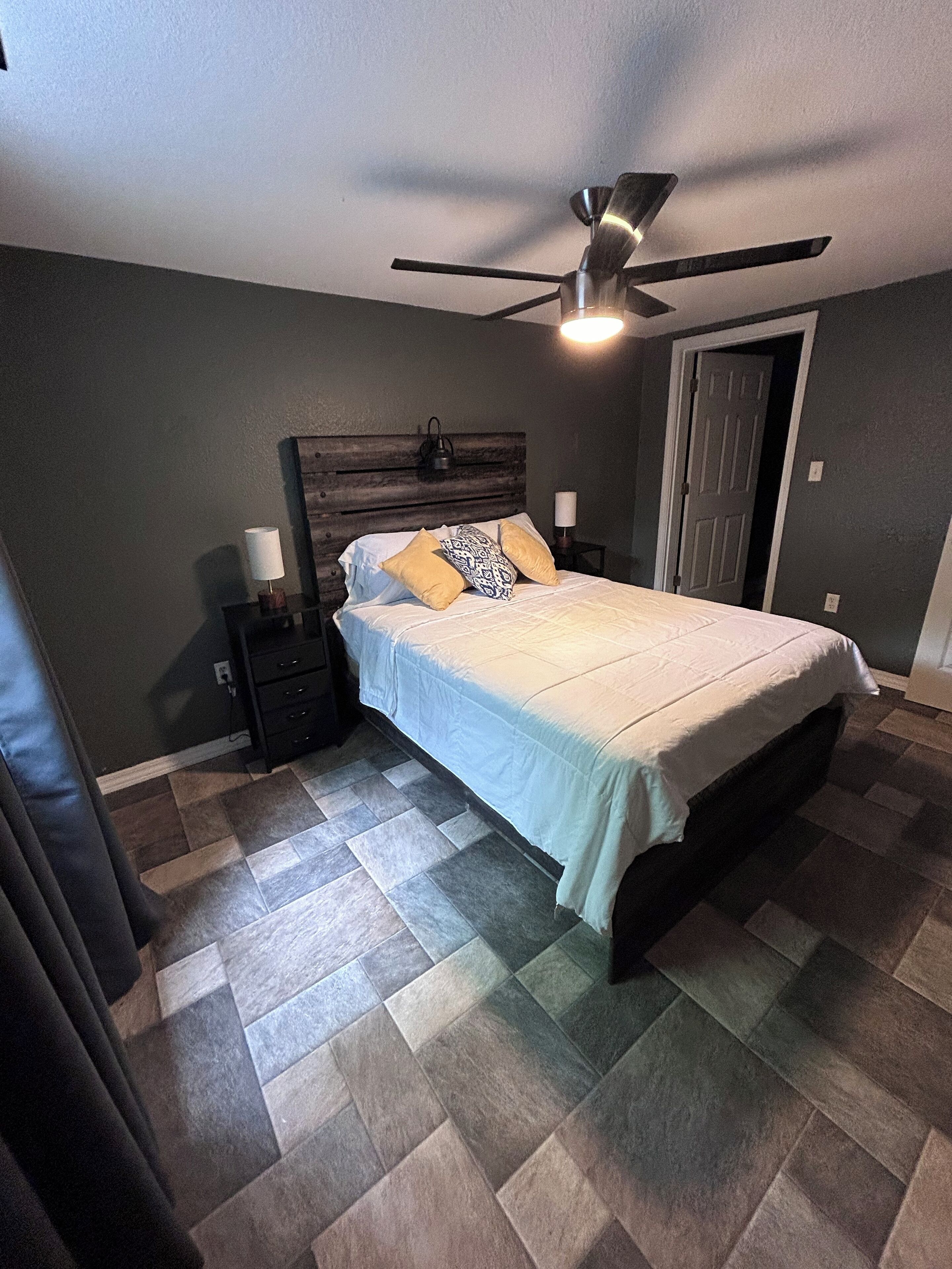 Een slaapkamer met een bed, nachtkastje, lamp, plafondventilator en een deur.