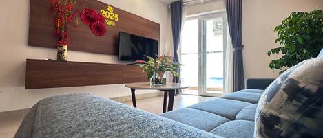 A modern living room with a sofa, a TV mounted on the wall, a small table with a plant, and a window with curtains.
