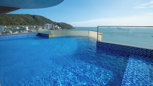 Una piscina con agua cristalina azul, una barandilla de vidrio y vistas al mar y a una colina con edificios.