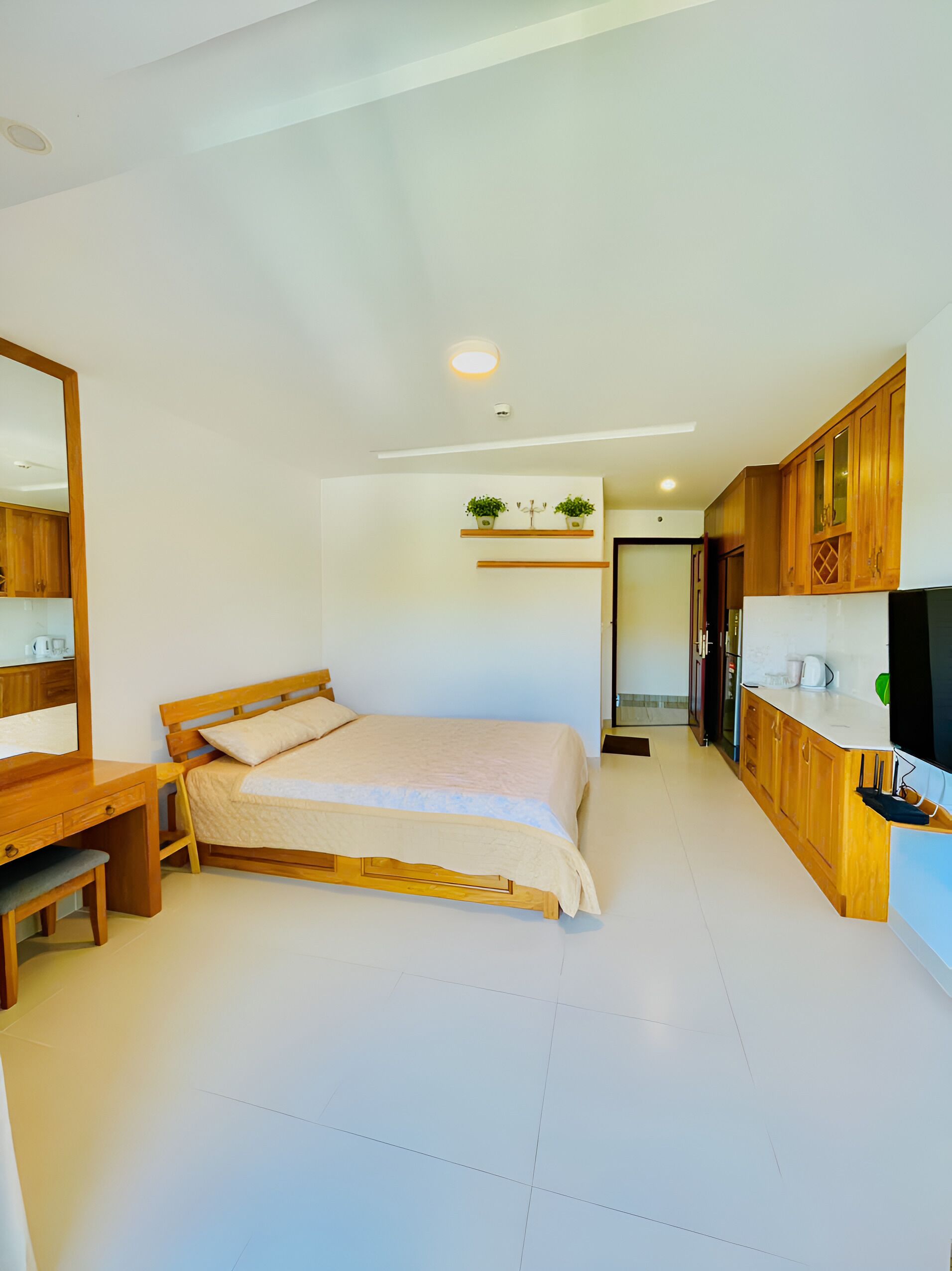 A bedroom with a wooden bed, a desk, a chair, a TV, and a kitchen area with wooden cabinets and a countertop.