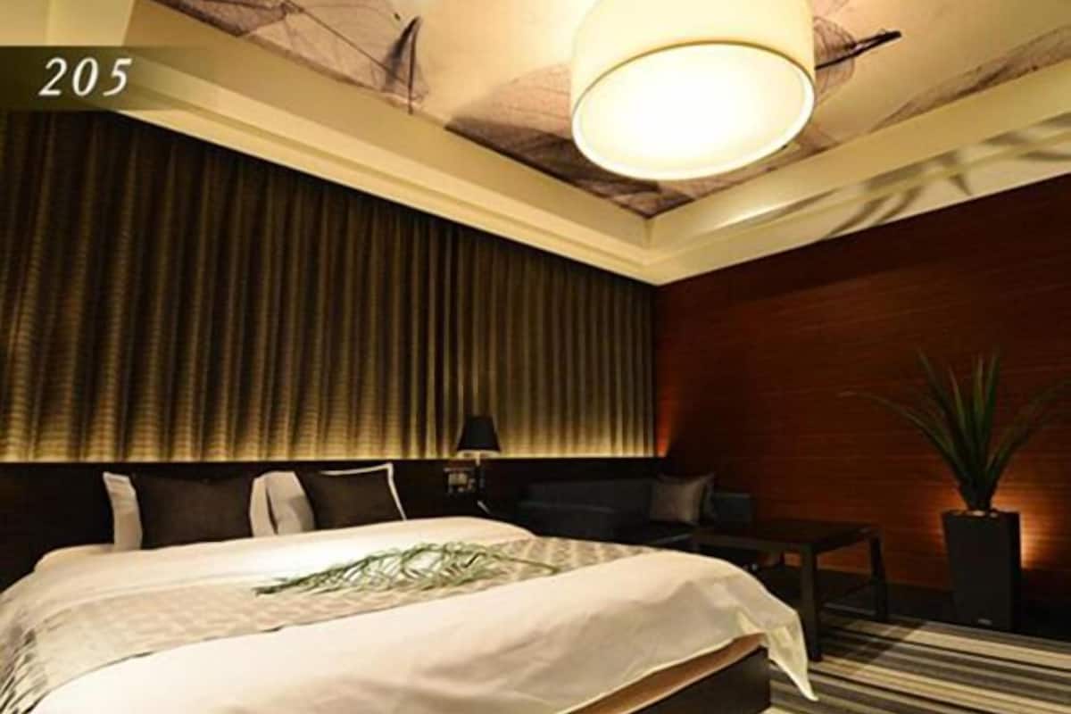 A hotel room with a large bed, a ceiling light, a potted plant, and a window with curtains.