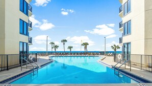 A swimming pool with a view of the ocean, surrounded by palm trees and a building with balconies.