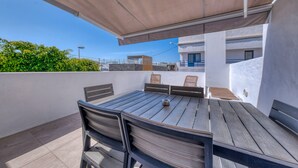 Een terras buiten met een houten tafel en stoelen, een overdekt gedeelte en uitzicht op een gebouw met balkons.