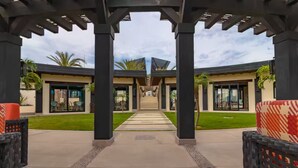 A modern outdoor pathway leading to a building with a covered entrance, surrounded by greenery and palm trees.