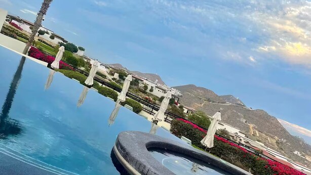 A clear view of a swimming pool with a curved edge, surrounded by a white fence and red flowers. In the background, there are buildings and a mountain.