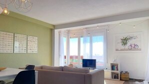 Una sala de estar con sofá, televisor y comedor con sillas. Hay cuadros en la pared y una estantería llena de libros.
