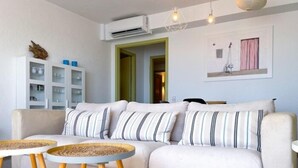 Una sala de estar con un sofá beige, cojines a rayas, dos mesas redondas con patas de madera y un aire acondicionado instalado en la pared.